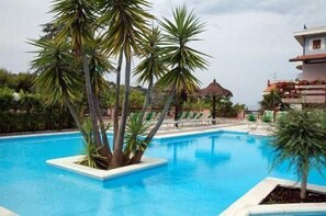 A swimming pool surrounded by palm trees and lounge chairs, with a building in the background. - Villaggio Verde Cupra (Cupra Marittima)