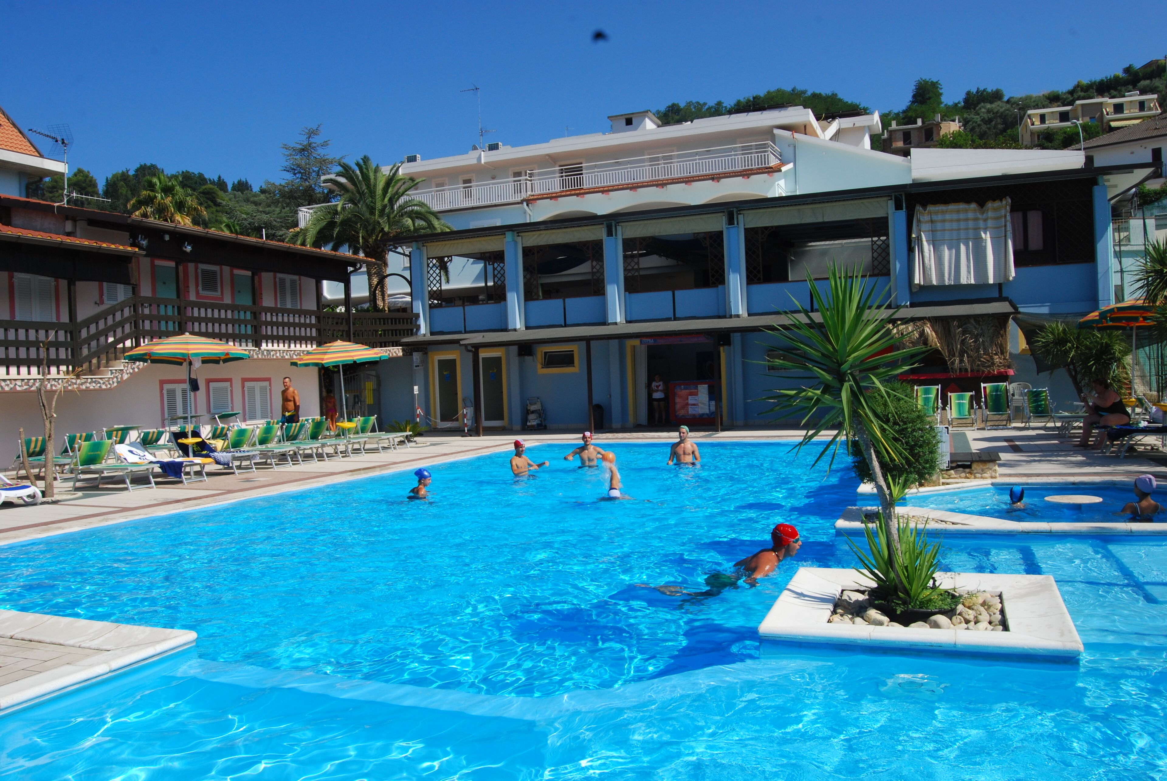 A swimming pool with lounge chairs and umbrellas, surrounded by buildings with balconies and a clear sky.