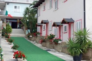Weiße Häuser mit rosa Fensterläden, ein grün bepflasterter Weg und Blumentöpfe.