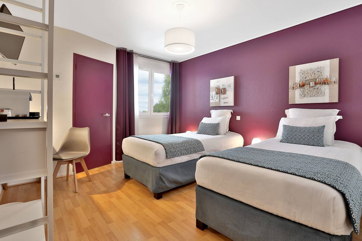 A hotel room with two beds, a window with curtains, a white chair, and a shelf with items.