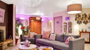 A modern living room with a grey sofa, a yellow armchair, and a coffee table. There is a fireplace, a clock, and a sign indicating the location of the bathrooms. - Hôtel Europa (Maussac)