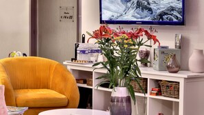 A modern living room with a large flat-screen TV, a unique chandelier, a vibrant yellow chair, and a small round table with a vase of flowers. - Hôtel Europa (Maussac)