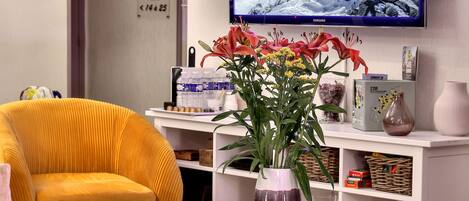 Una sala de estar moderna con un televisor de pantalla plana grande, una lámpara de araña original, un sillón amarillo vibrante y una mesita redonda con un jarrón de flores.