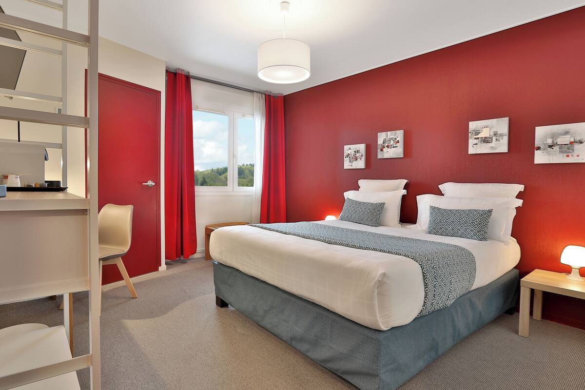 A bedroom with a large bed, a desk, and a chair. There are three paintings on the wall and a window with red curtains.