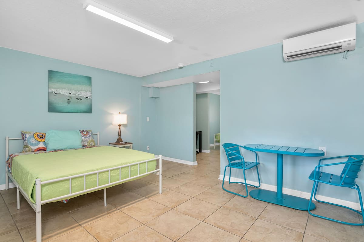 A room with a bed, a table with chairs, a wall-mounted air conditioner, and a painting.