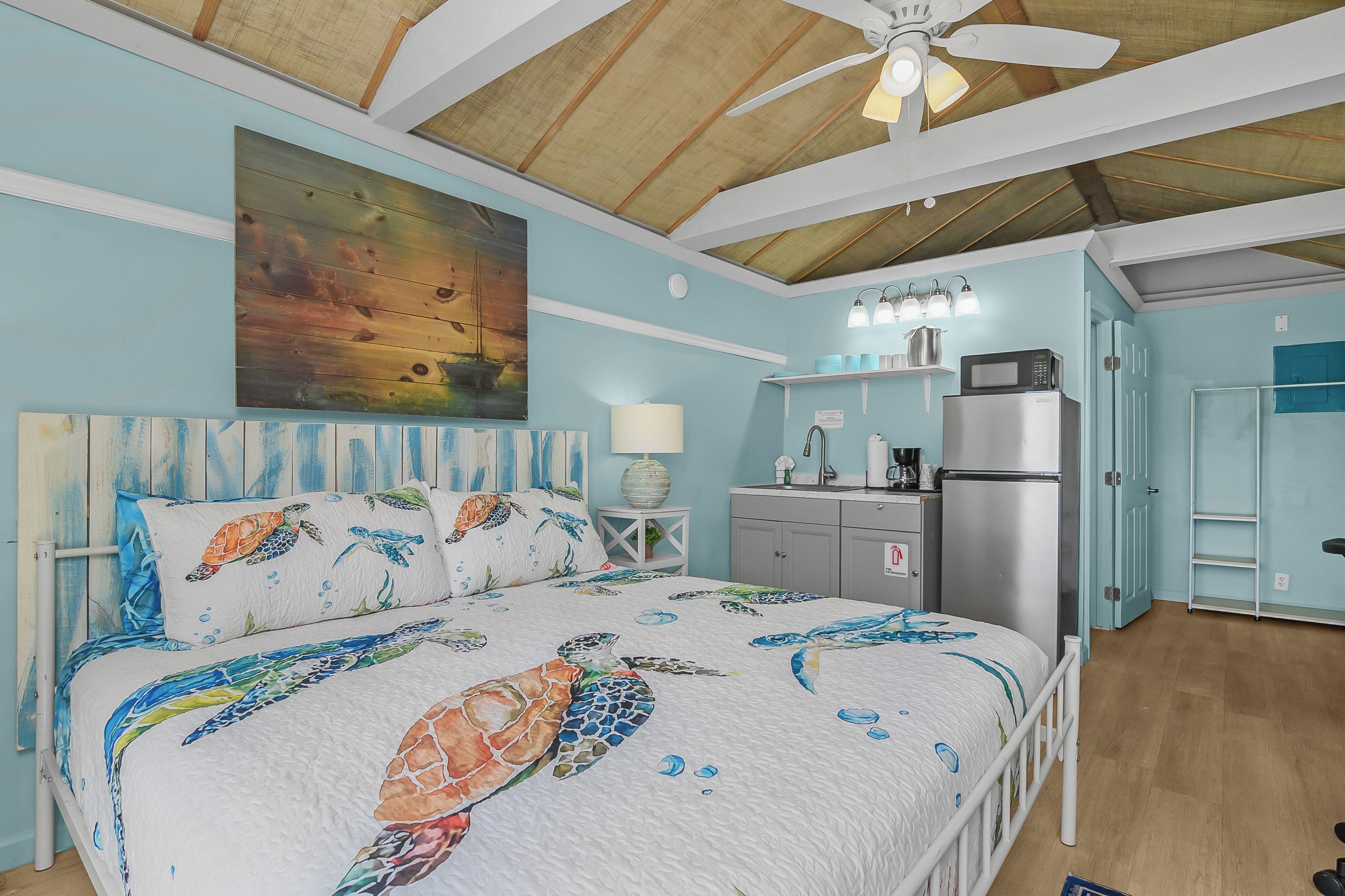 A bedroom with a bed, a ceiling fan, a painting, a kitchen area with a refrigerator, and a bathroom with a door.