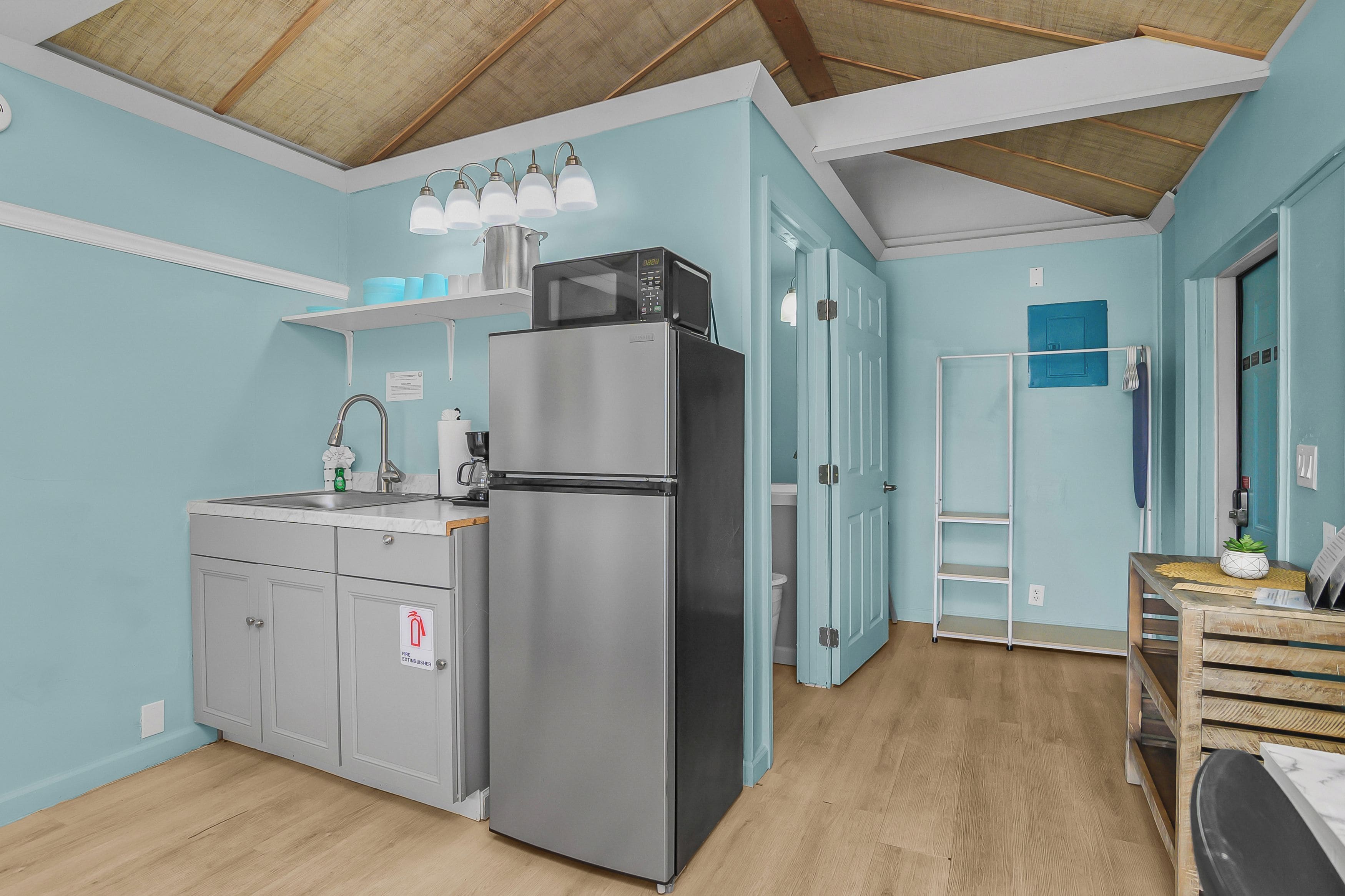 A compact kitchen with a stainless steel refrigerator, a sink, and a microwave. There is a shelf above the sink and a door leading to a bathroom.