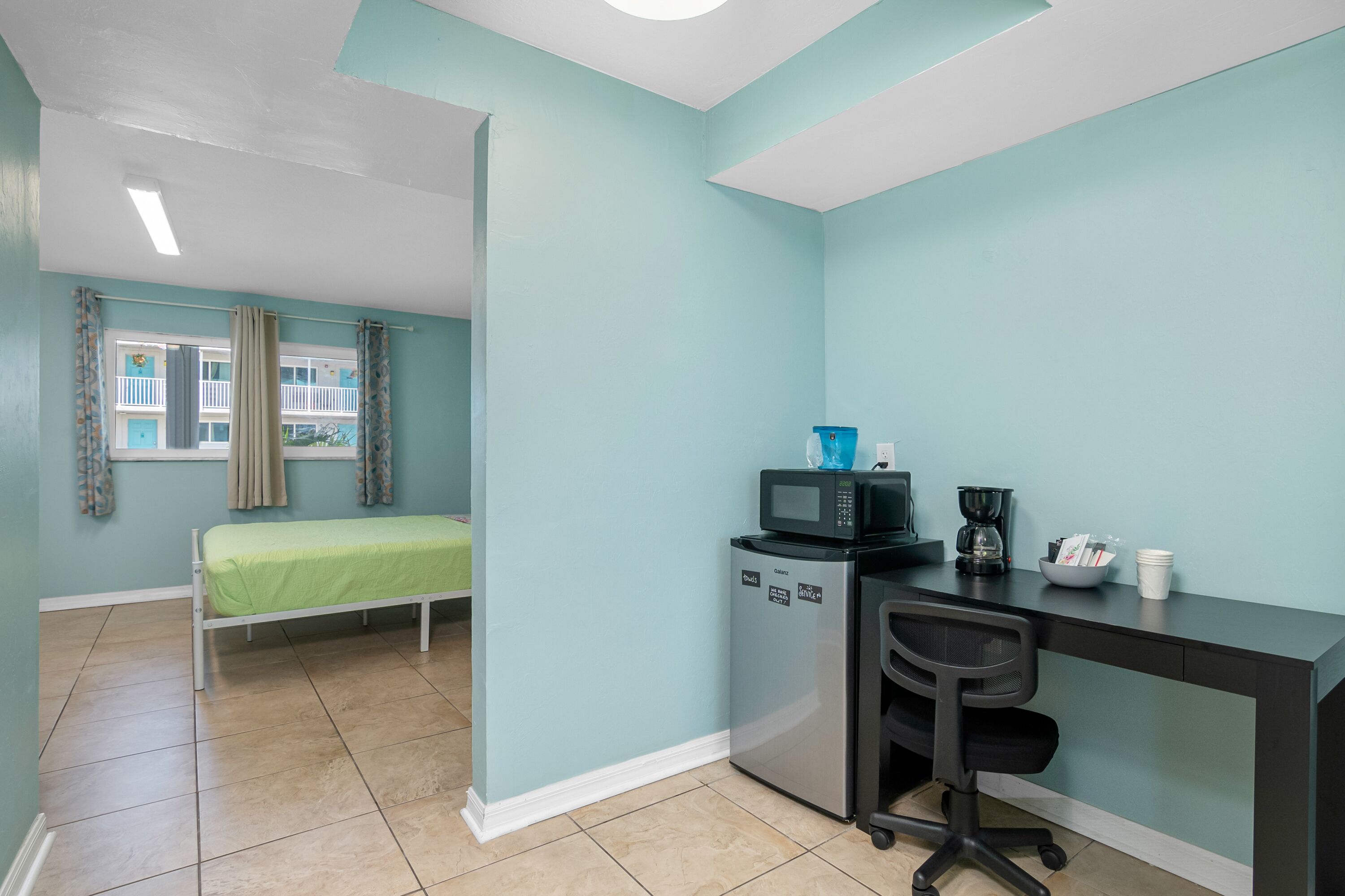 Une pièce compacte aux murs de couleur turquoise, comprenant un lit, une kitchenette équipée d’un four à micro-ondes, d’une machine à café et d’un petit réfrigérateur, ainsi qu’un bureau avec une chaise.