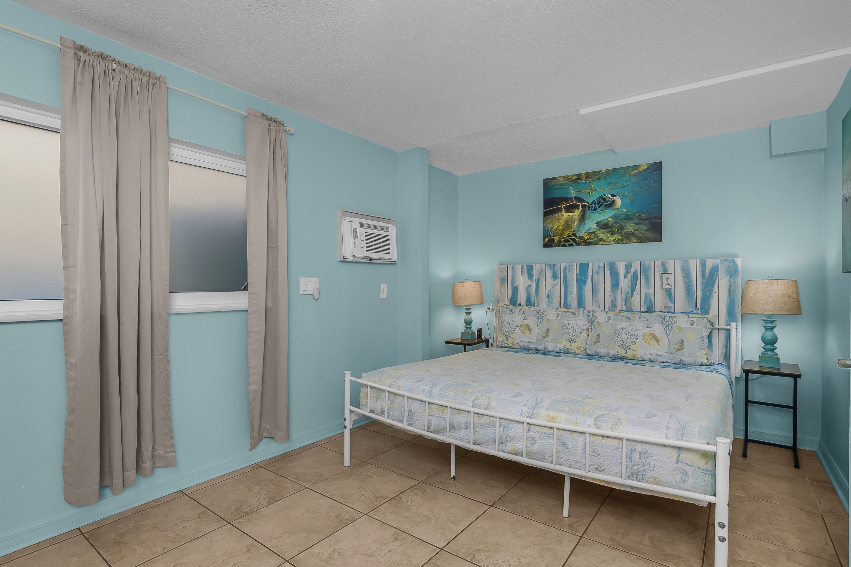 A bedroom with a single bed, a window with curtains, a wall-mounted air conditioner, a painting, and a bedside table with a lamp.