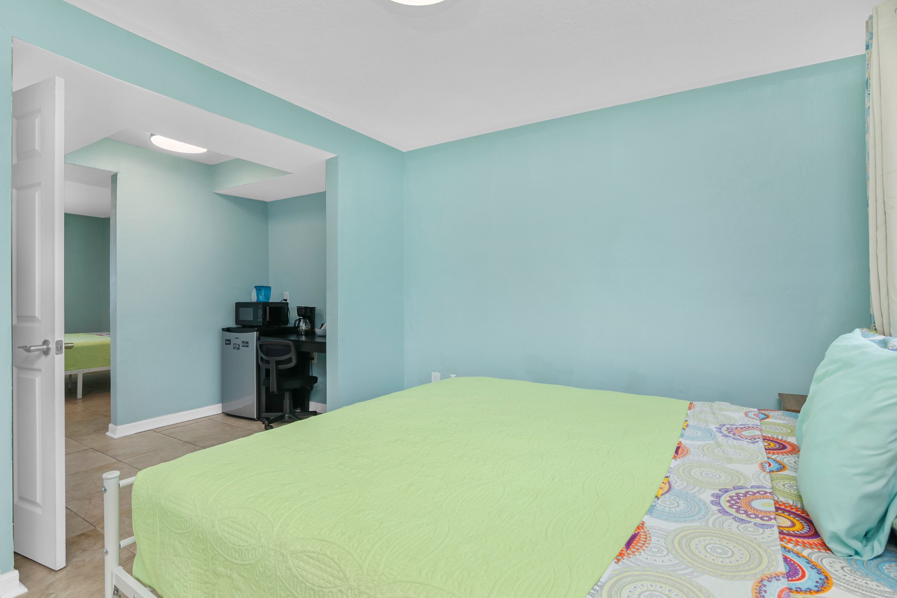 A bedroom with a bed, a desk with a computer, and a door leading to another room.