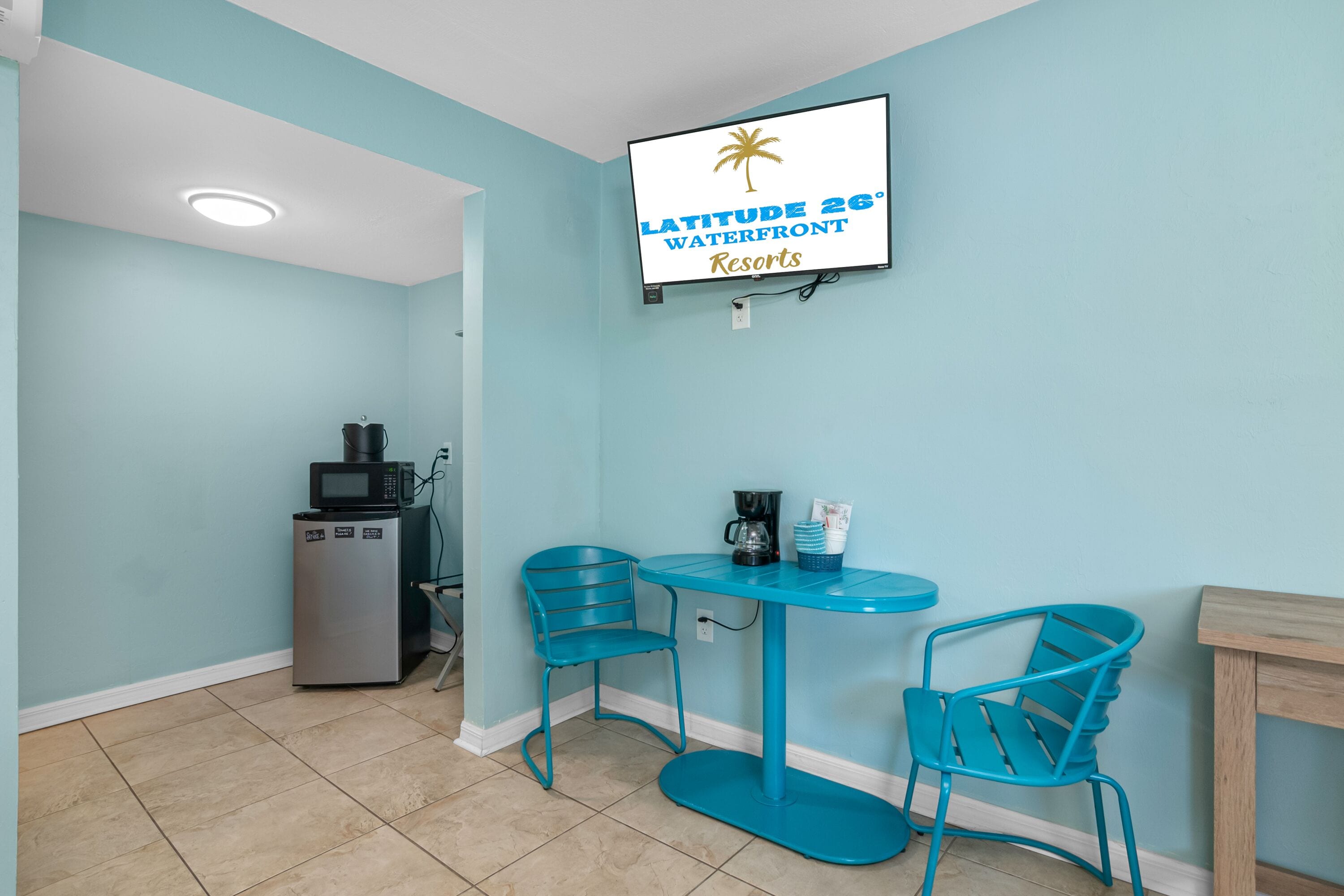 A room with a teal color scheme, a television mounted on the wall displaying 'LATITUDE 26° WATERFRONT RESORTS', a small refrigerator, a coffee maker, and two chairs around a table.