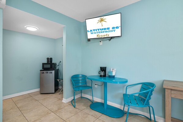 Een kamer met een turquoise kleurenpalet, een televisie aan de muur met daarop 'LATITUDE 26° WATERFRONT RESORTS', een kleine koelkast, een koffiezetapparaat en twee stoelen rond een tafel.