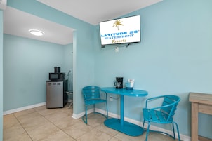 A room with a teal color scheme, a television mounted on the wall displaying 'LATITUDE 26° WATERFRONT RESORTS', a small refrigerator, a coffee maker, and two chairs around a table.