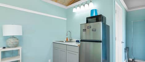 A kitchen with light blue walls, a stainless steel refrigerator, a sink, and a white shelf with a lamp.
