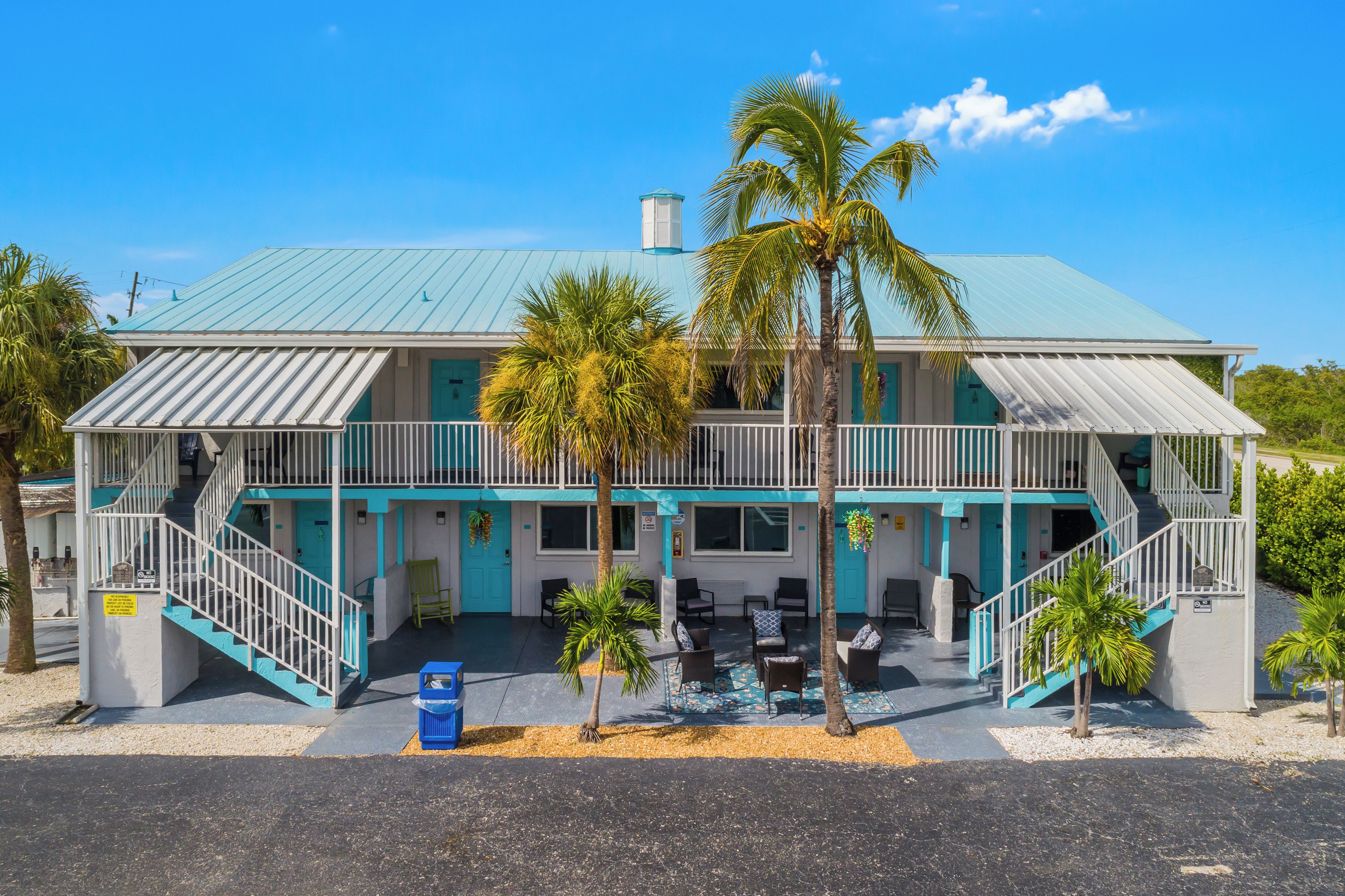 A two-story building with a blue roof, a balcony, and a small outdoor seating area with chairs and tables.