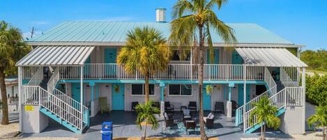 A two-story building with a blue roof, a balcony, and a small outdoor seating area with chairs and tables.