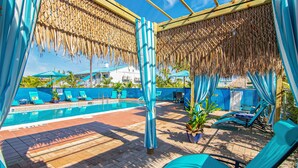 Un área junto a la piscina con sillones de descanso, una piscina y una zona de estar cubierta con techo de paja.