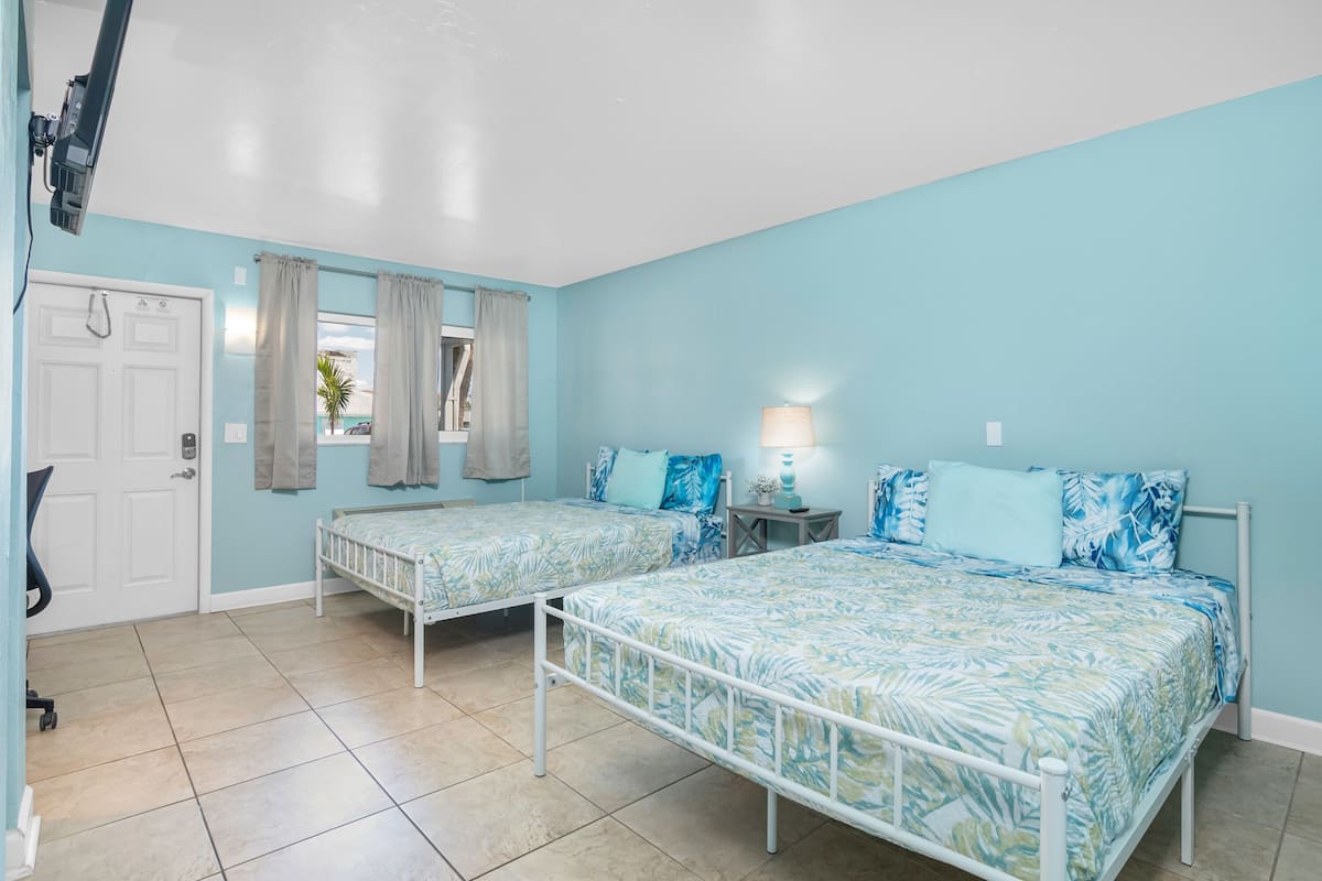 A room with two single beds, a white door, a window with curtains, a small table with a lamp, and a ceiling fan.