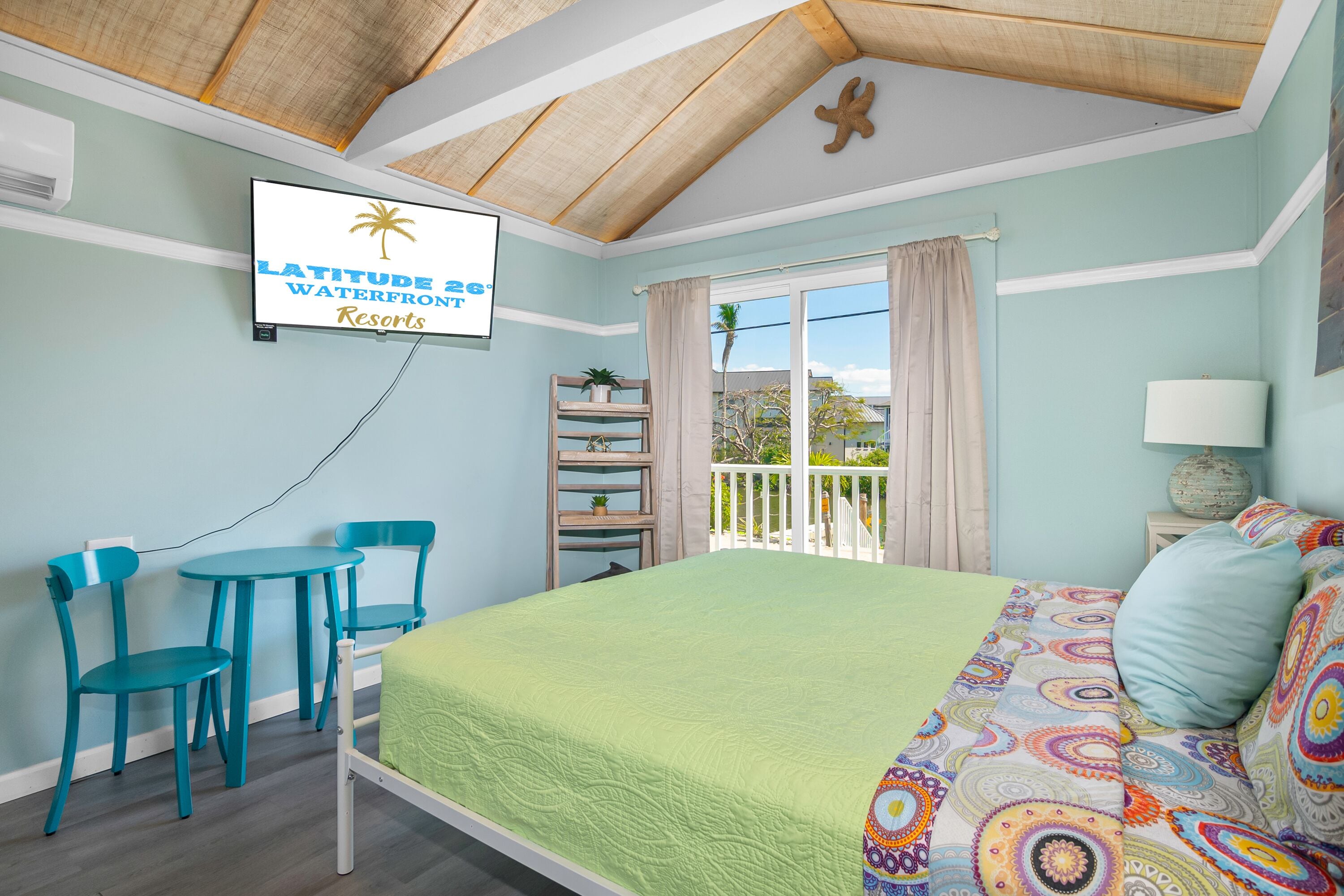 A bedroom with a bed, a desk, and chairs, a TV on the wall, and a balcony with a view.