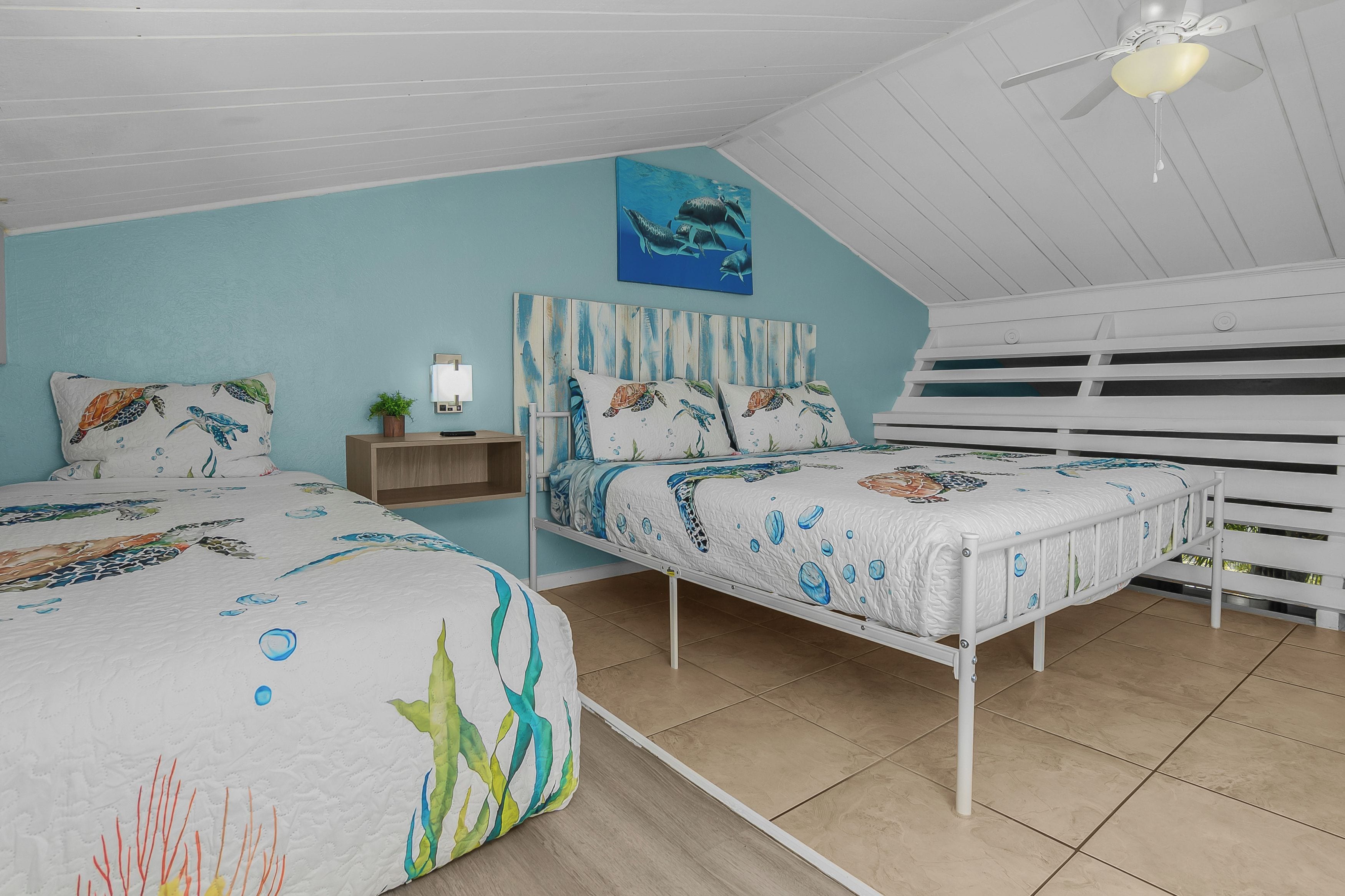 Un dormitorio con dos camas, ventilador de techo, un cuadro y una mesita de noche.