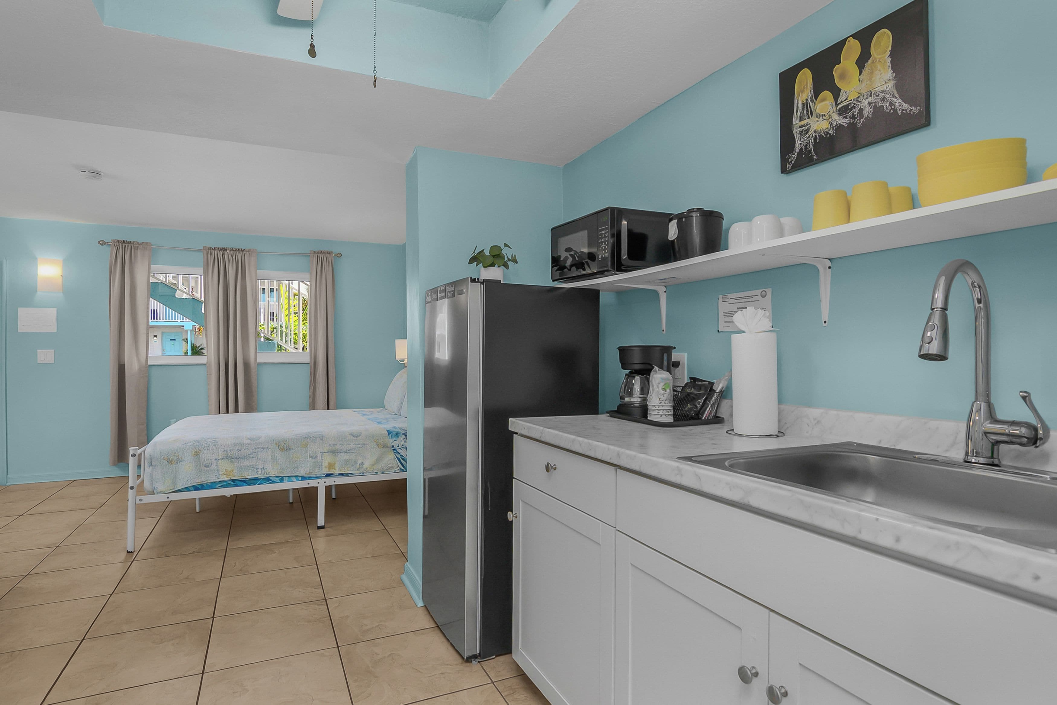 A compact kitchen with a refrigerator, microwave, and sink. There is a bed with a window view and a small plant on the counter.