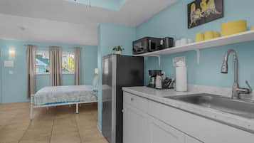 A compact kitchen with a refrigerator, microwave, and sink. There is a bed with a window view and a small plant on the counter.