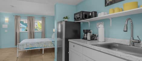 A compact kitchen with a refrigerator, microwave, and sink. There is a bed with a window view and a small plant on the counter.