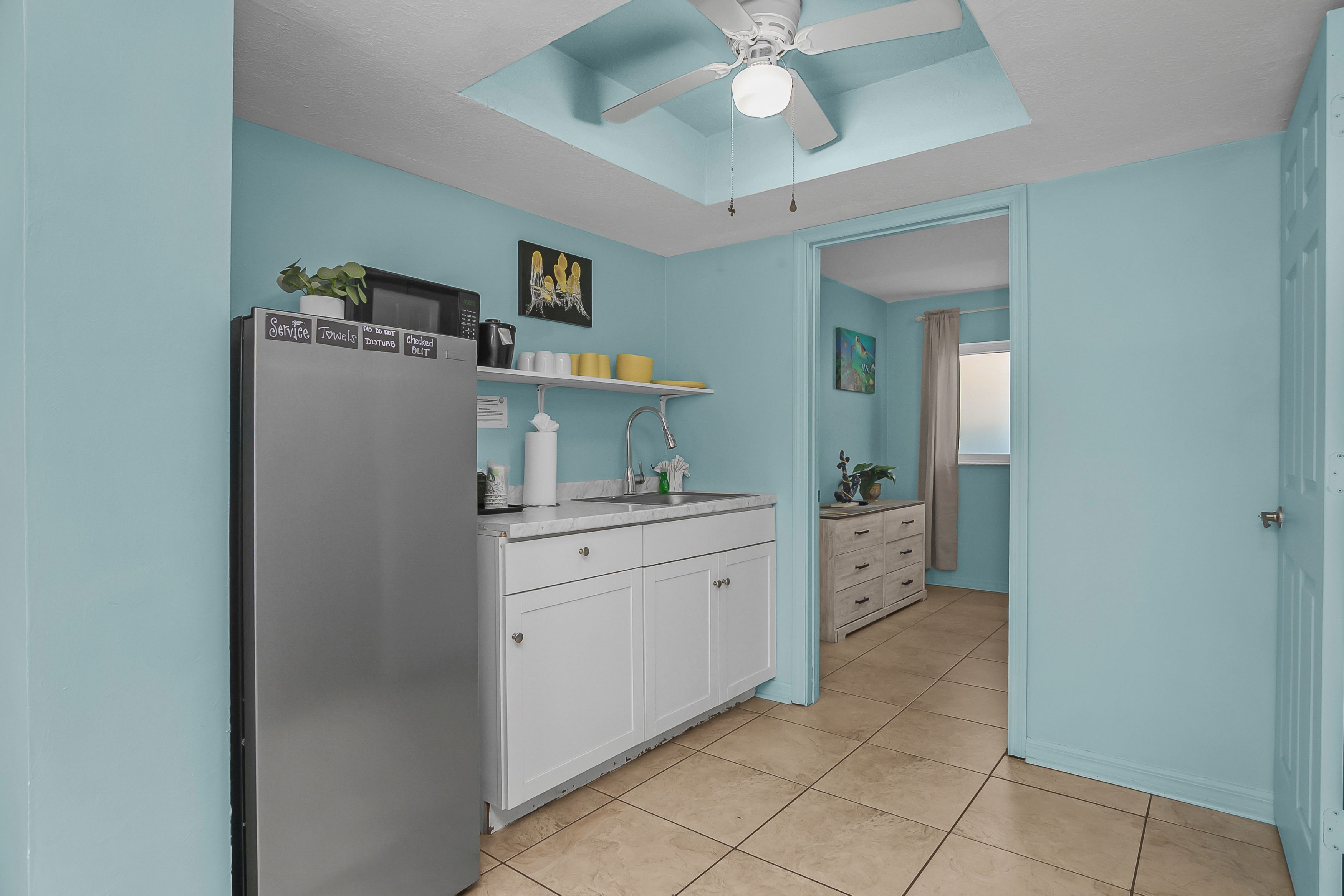 Cocina equipada con refrigerador, pileta y alacenas. Cuenta con un ventilador de techo y una ventana con cortinas.