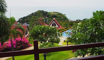 Vista de un resort con piscina rodeada de exuberante follaje y flores coloridas. Varias edificaciones con techos rojos y una colina al fondo.