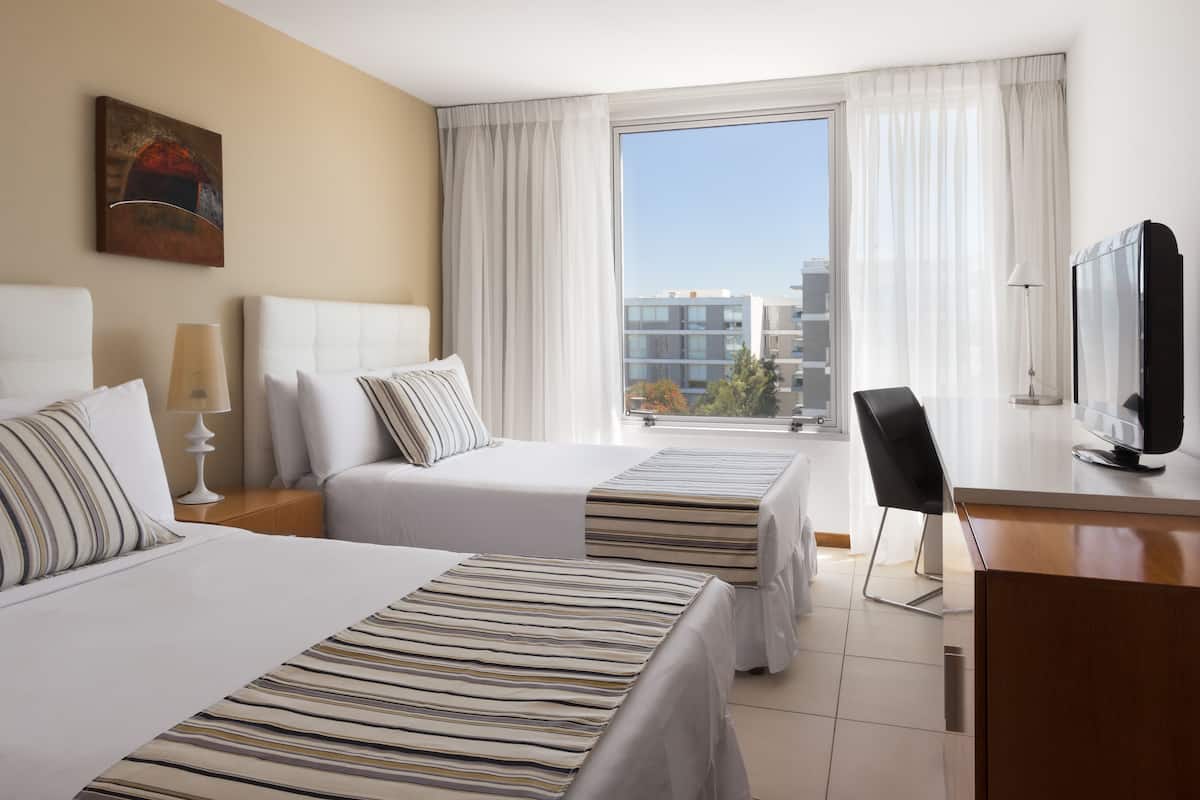 Habitación de hotel con dos camas, un escritorio, una silla, un televisor y una ventana con vista a la ciudad.