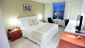 A hotel room with a bed, bedside table, desk, chair, and a view of the outside. - Real Colonia Hotel & Suites (Colonia del Sacramento)