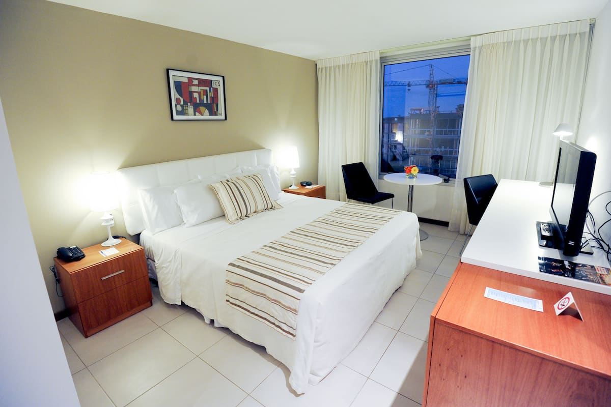 Habitación de hotel con cama, mesita de noche, escritorio, silla y vistas al exterior.