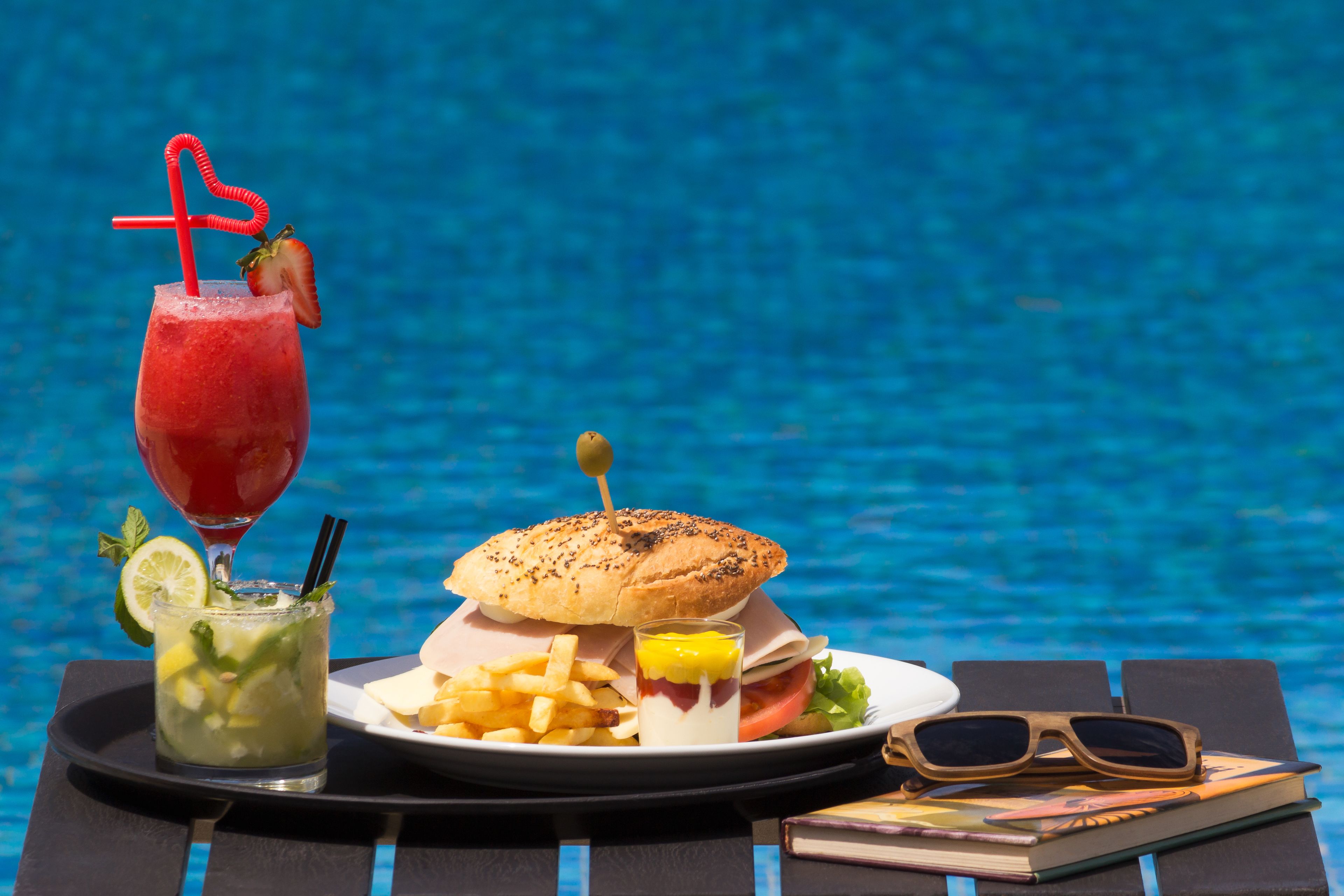 Comida junto a la piscina, con una hamburguesa, papas fritas, una bebida y gafas de sol.