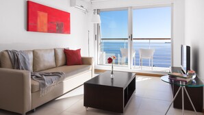A modern living room with a sofa, coffee table, and a glass table with books. A red painting is on the wall, and there is a view of the sea through large windows. - Real Colonia Hotel & Suites (Colonia del Sacramento)