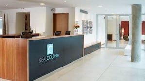 A reception desk with a logo, a modern interior with a glass door, and a pillar. - Real Colonia Hotel & Suites (Colonia del Sacramento)