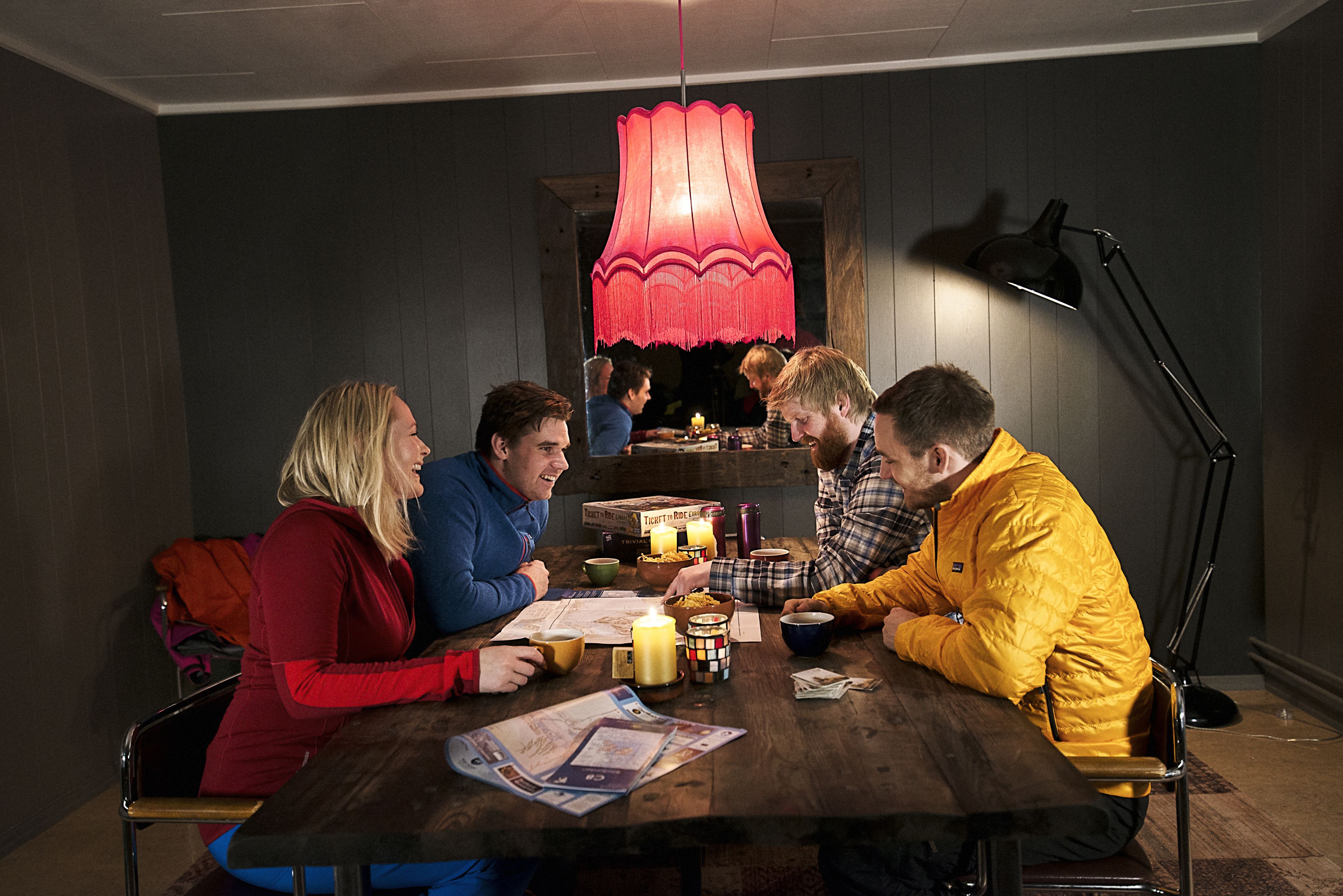 Een groep mensen zit rond een houten tafel in een kamer met donkere wanden en een rode hanglamp.