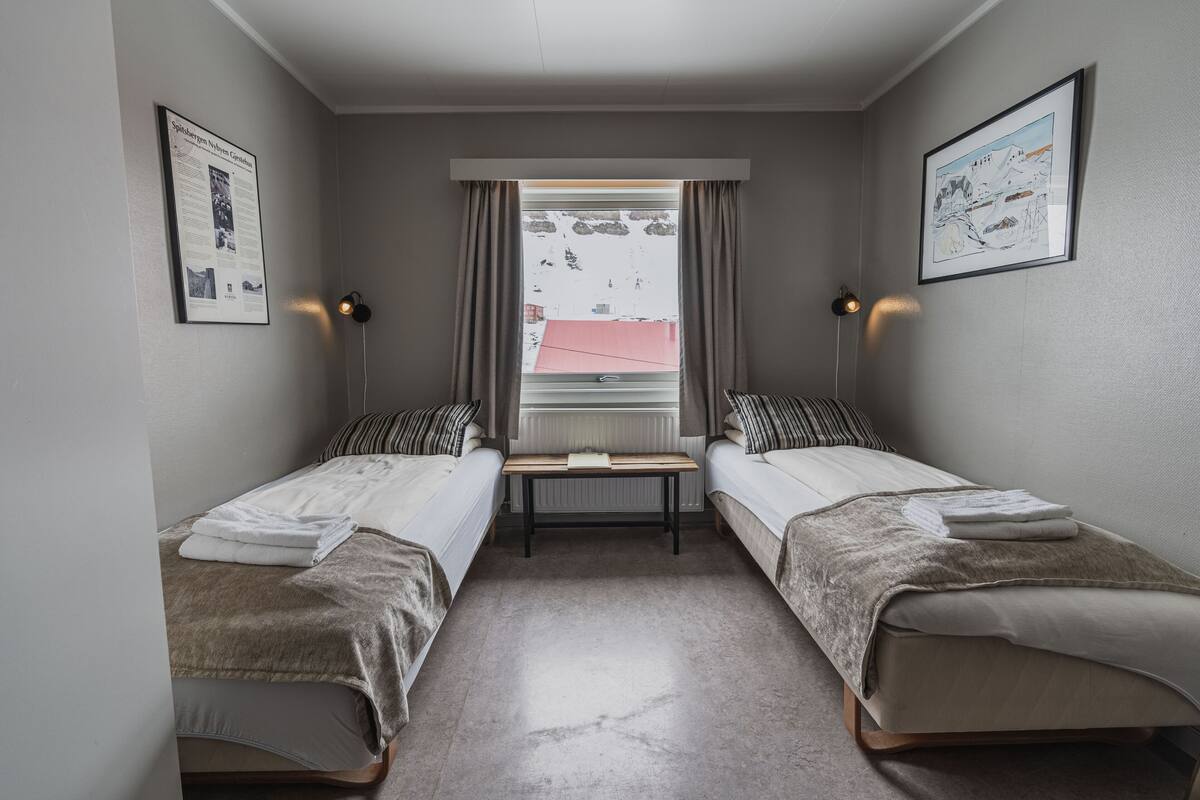 A small, well-lit room with two single beds, a window with curtains, and framed pictures on the walls.