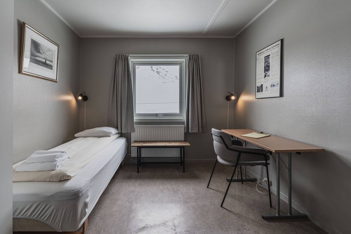 A small, neatly arranged room with a single bed, a desk, and a chair. There is a window with curtains, a radiator, and two wall-mounted lamps.
