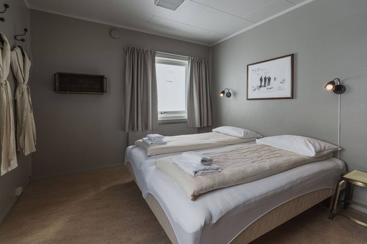 A hotel room with two beds, a window with curtains, a framed picture on the wall, and a bedside table with a lamp.