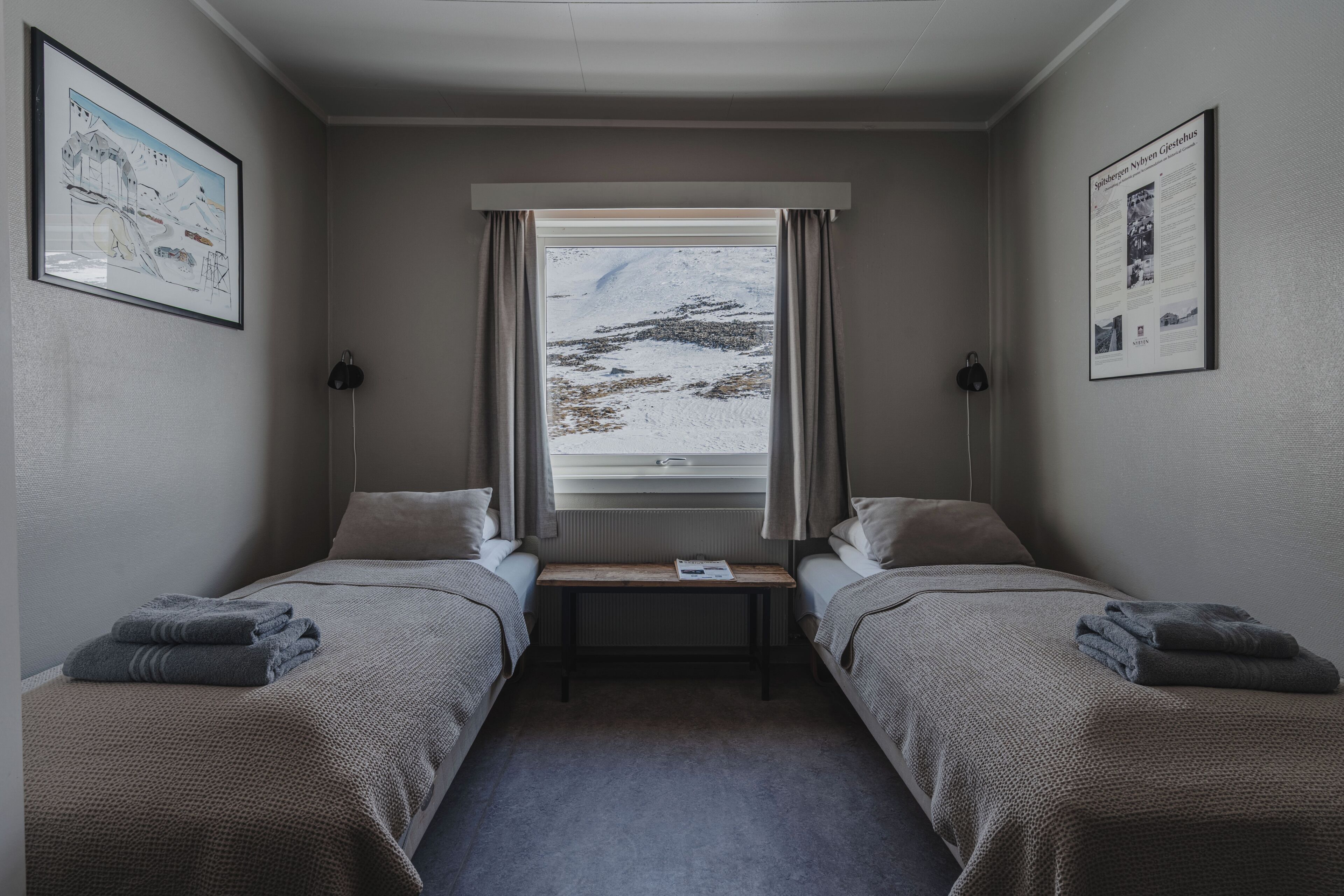 A hotel room with two single beds, a small table, a window with a view of a snowy landscape, and framed pictures on the walls.