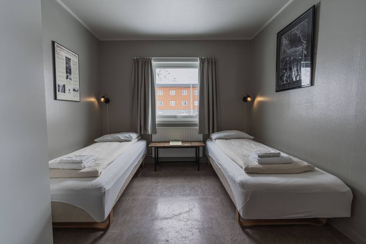 A small, clean room with two single beds, a small table, and a window with curtains.