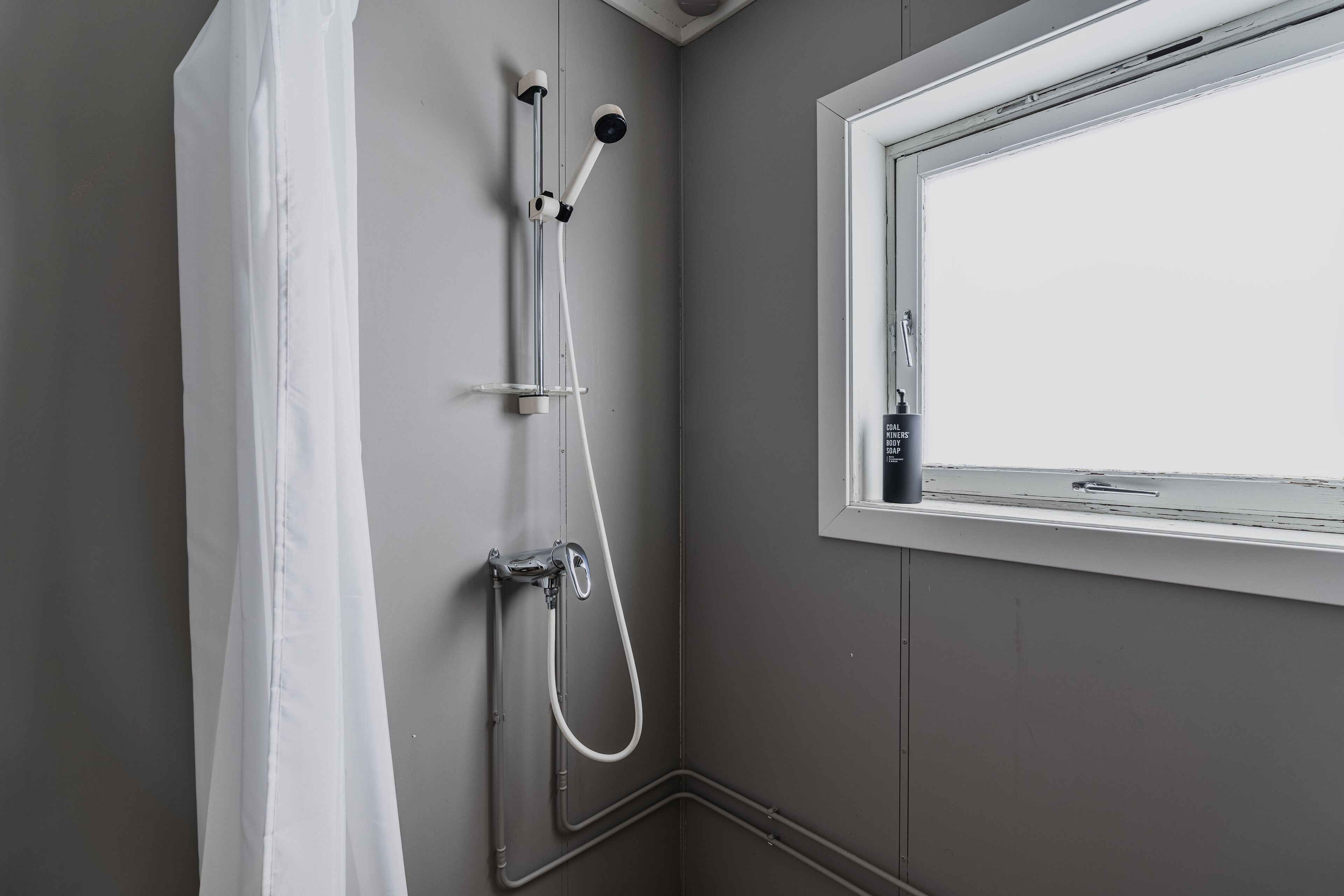 Ducha con cabezal de ducha de mano y grifo de pared, ventana con vistas y cortina de ducha blanca.