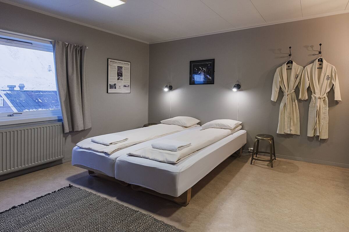 A bedroom with a bed, two bedside tables, a window with curtains, a radiator, a wall-mounted lamp, and two robes hanging on hooks.