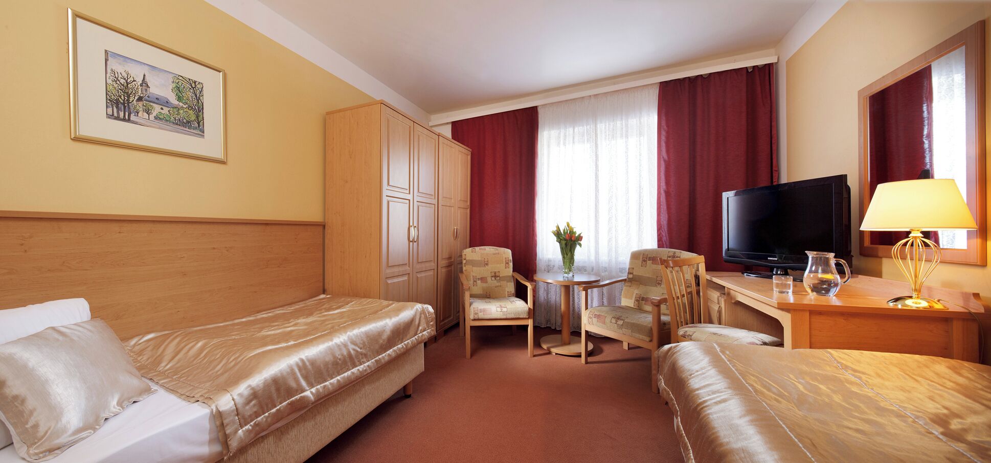 A hotel room with two beds, a desk with a TV, a chair, a small table with a vase, and a window with red curtains.