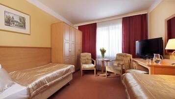 A hotel room with two beds, a desk with a TV, a chair, a small table with a vase, and a window with red curtains.