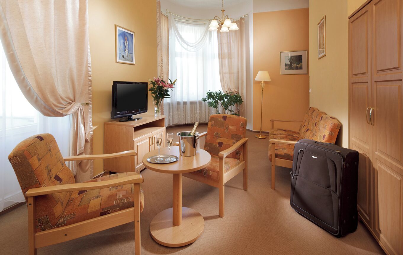 A hotel room with a wooden dining table, two patterned chairs, a television, a minibar, a suitcase, and a large window with sheer curtains.