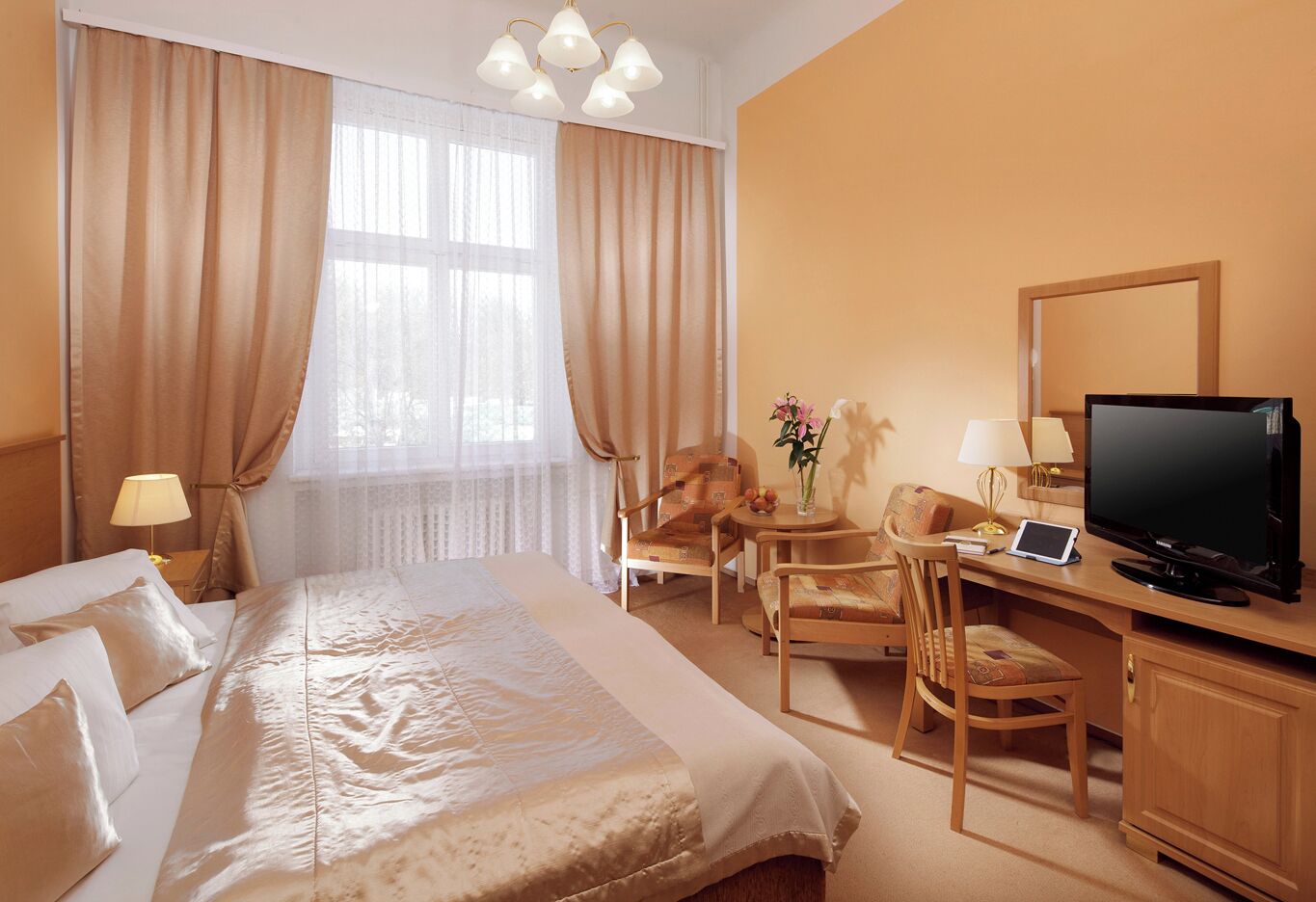 A hotel room with a large bed, a desk with a television, a chair, and a small table with a vase of flowers.