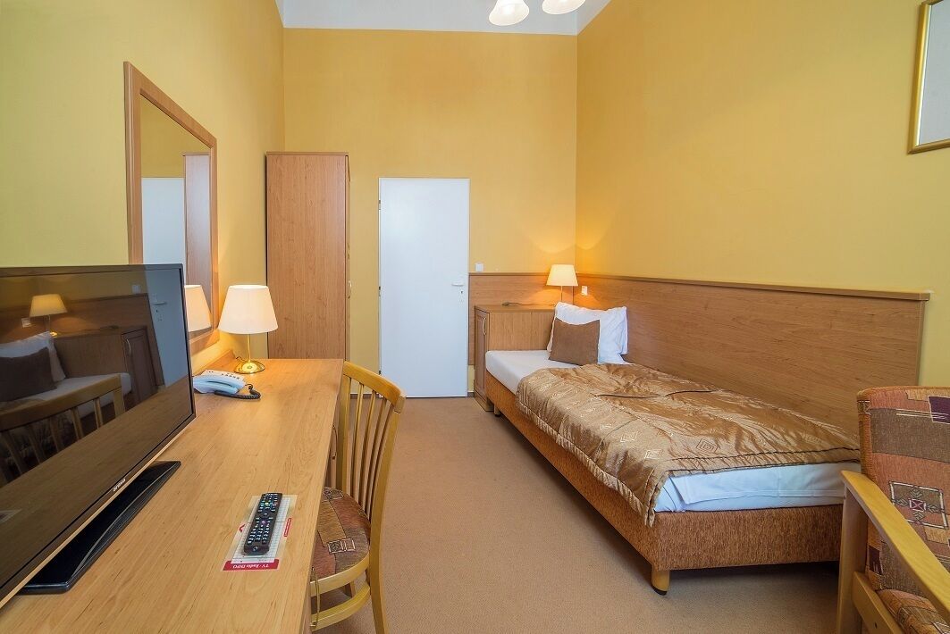 A hotel room with a bed, a desk with a TV, a chair, a lamp, and a bedside table.