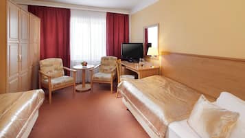 A hotel room with two beds, a desk with a TV, a small table with chairs, and a window with curtains.