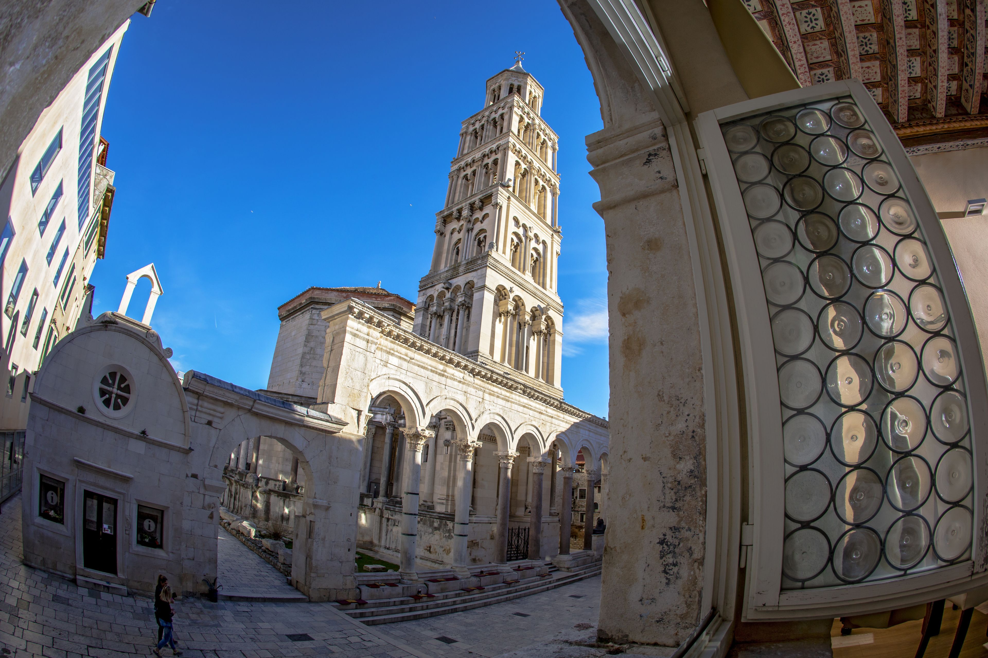 A historic building with a prominent tower, arched walkways, and a clear blue sky.